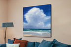 Canvas print of a beach scene with a blue sky and clouds, hanging on a beige wall above a sofa.