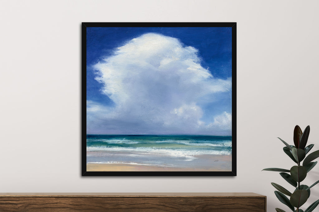 Framed artwork of a beach scene with blue sky and clouds on a white wall.
