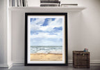 Framed beach print on a wall next to a shelf with books and a basket.